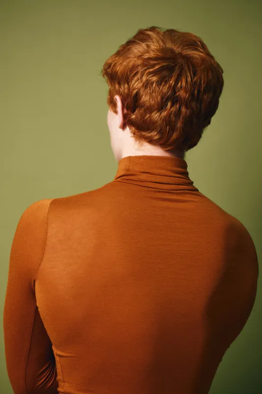 Person with red hair seen from behind wearing a brown turtleneck sweater against green background. Portrait style emphasizing hair texture and clothing details.