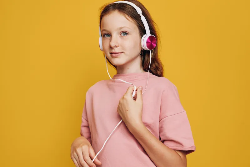 Young girl with long hair wearing pink headphones and a pink sweater stands against a yellow background. She looks confident and relaxed.