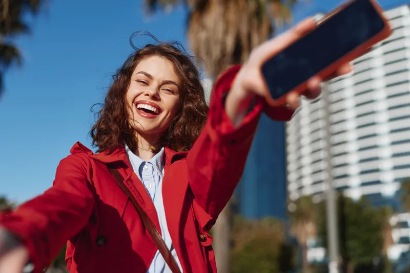Young woman with curly hair selfies outdoors in a red jacket, smiling brightly against a sunny city backdrop.