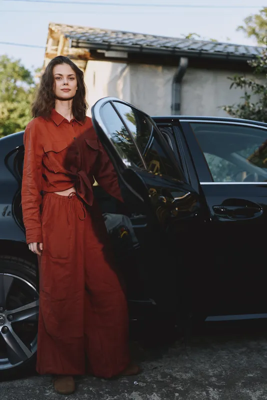 Confident woman in a rust red outfit stands beside a sleek car outdoors, sunlight highlights fabric texture, relaxed lifestyle moment, casual chic, natural colors, urban backdrop