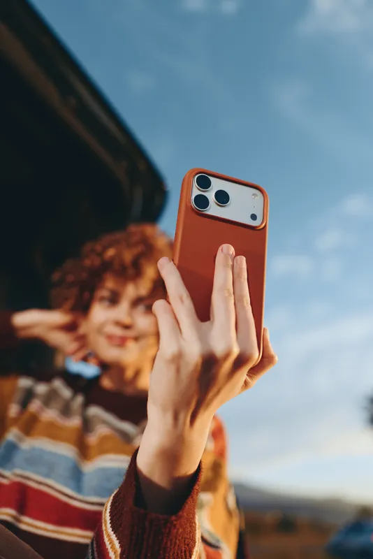 Woman lifestyle smartphone selfie outdoors smiling curly hair in background, close up hands holding red phone, knit sleeve visible, blue sky and soft bokeh, casual travel moment today