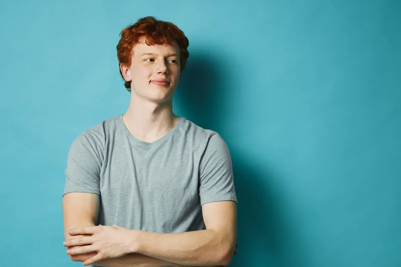 Young man with red hair and light freckles wearing a gray t shirt standing with arms crossed looking away on blue background studio portrait.