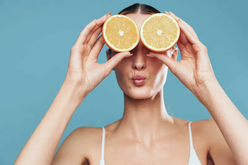 Woman holds orange citrus slices over eyes on blue background, showing skincare beauty, spa wellness, fresh vitamin health concept, closeup portrait of female hands with fruit