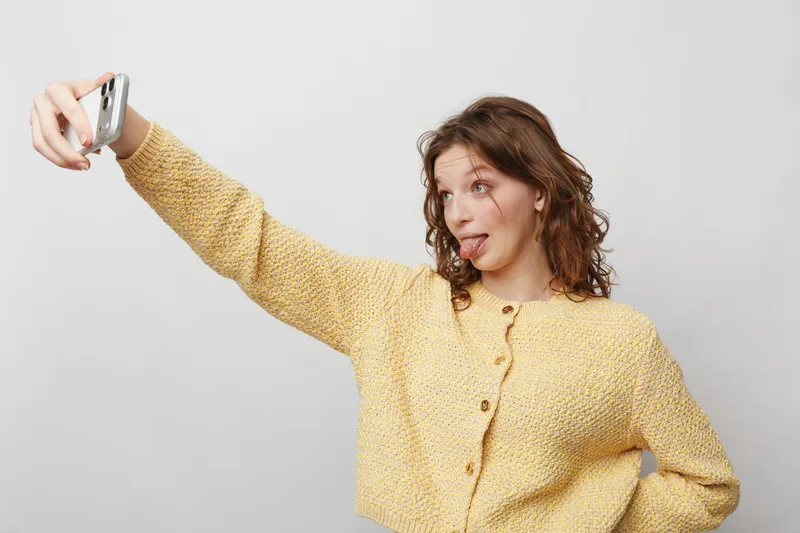 Young woman in yellow sweater takes a selfie with a smartphone, making a playful face and sticking out her tongue. Casual style, isolated on white background, concept of fun and youth.