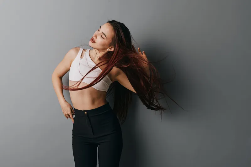 Young woman with long hair wearing white crop top and black pants posing against gray background in studio. Female model showing movement with closed eyes and casual style.