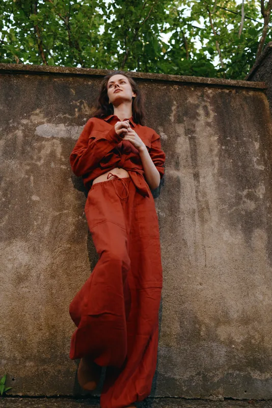 A stylish young woman in a red outfit leans against a weathered wall outdoors, looking upward with a calm, confident expression in a relaxed urban moment