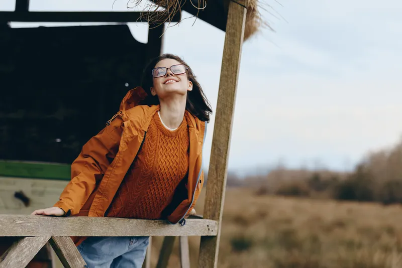 Smiling woman outdoors in autumn field, leaning on wooden structure, wearing orange sweater and glasses, relaxed lifestyle portrait, nature background, natural light, warm mood