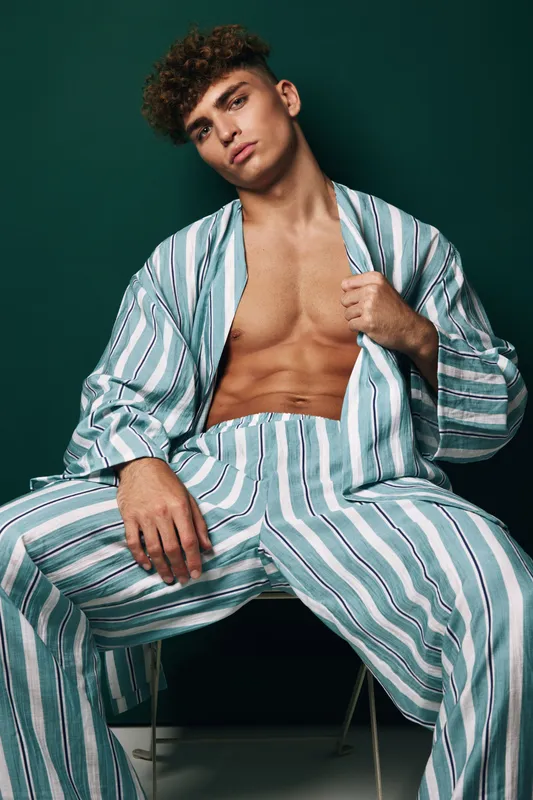 Young man with curly hair sitting in striped loose pants and open shirt showing toned chest against dark green background. Studio portrait of casual adult fashion male model with relaxed pose.