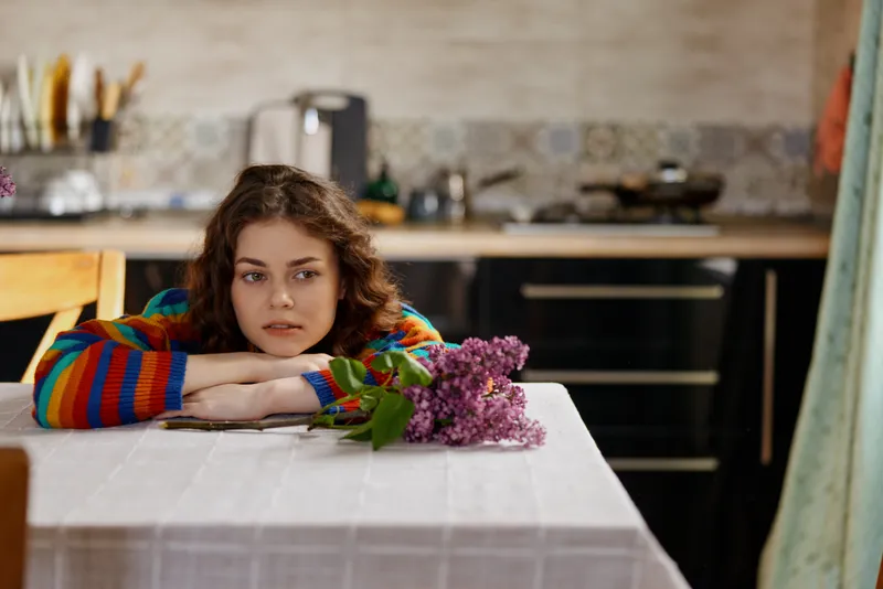 Calm woman resting at a dining table in a cozy kitchen, flowers and bouquet nearby, capturing a quiet home lifestyle moment with soft light and reflective mood.