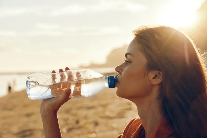 Woman drinking water outdoors at sunset holding a clear plastic bottle side profile with focused expression warm sunlight on face dry landscape background healthy lifestyle hydration time
