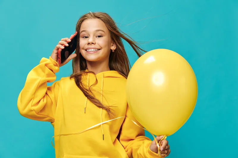 Girl in yellow hoodie holding yellow balloon and talking on smartphone with smile, standing against blue background. Child with long hair and happy face in casual clothes.