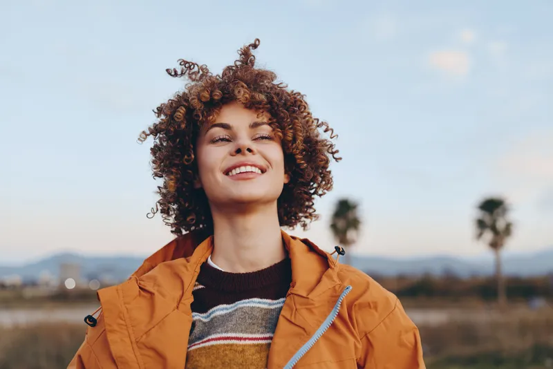 Woman with curly hair outdoors, smiling in warm sunshine, wearing orange jacket, relaxed lifestyle portrait, light breeze, soft sky background, golden hour, cheerful mood