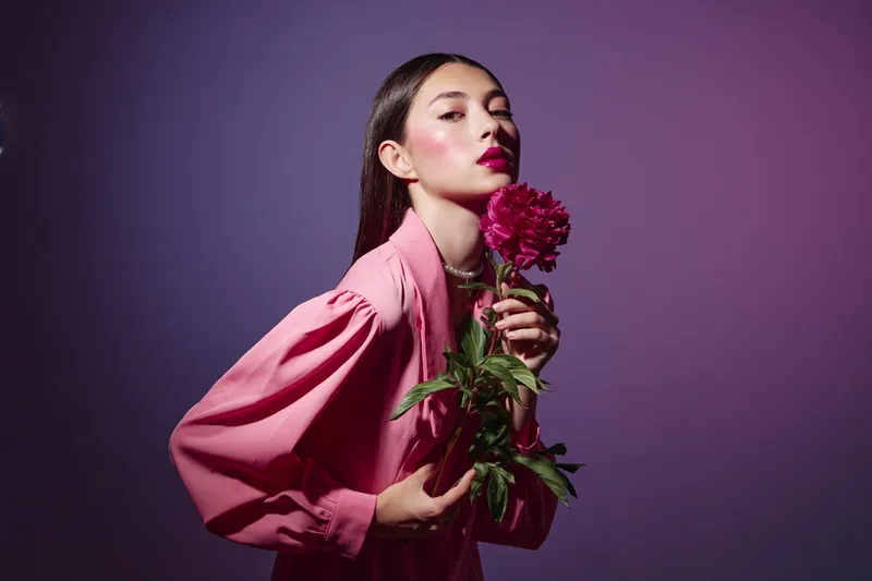Young woman in pink fashion with bold makeup holding a bright red flower close to her face. Studio portrait on pastel violet background. Concept of beauty and creative fashion.