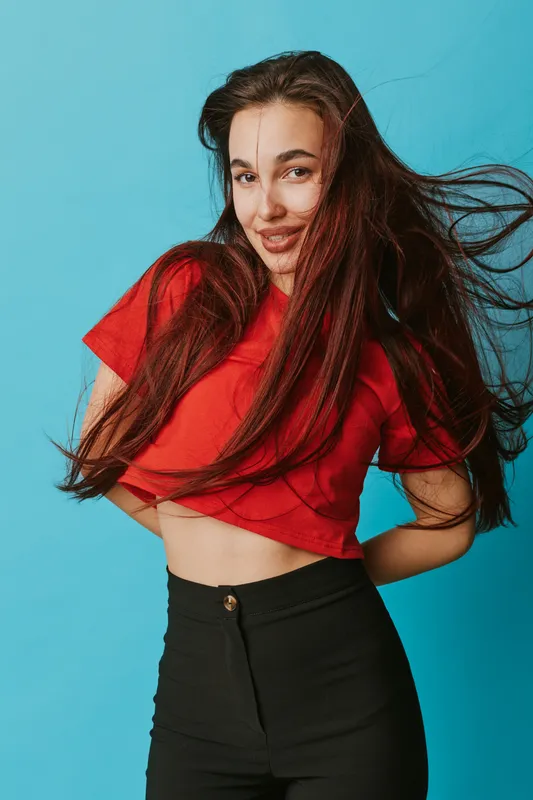 Young woman with long hair smiling and posing in red crop top and black pants against blue background. Casual style, confident expression and natural beauty with flowing hair movement.