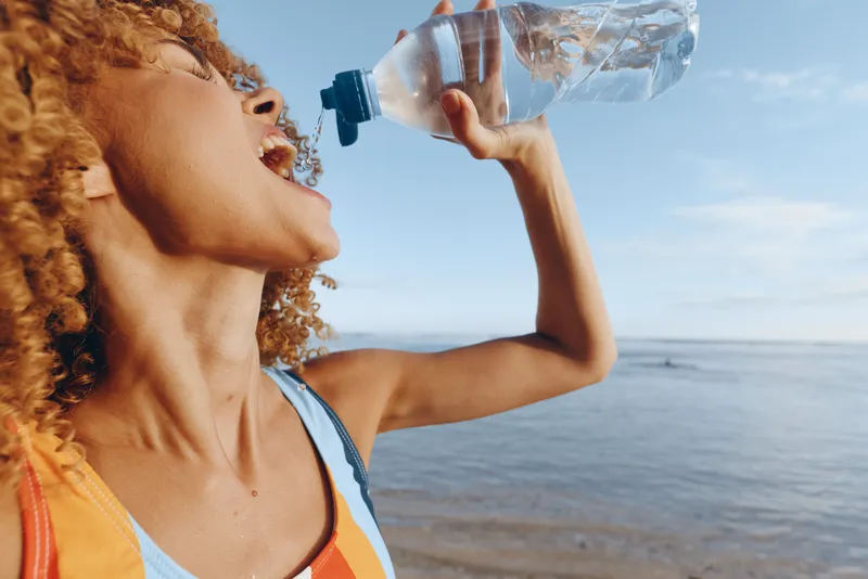 Young woman with curly hair drinks water from plastic bottle outdoors near the sea on sunny day, refreshing moment, casual clothing, clear blue sky background