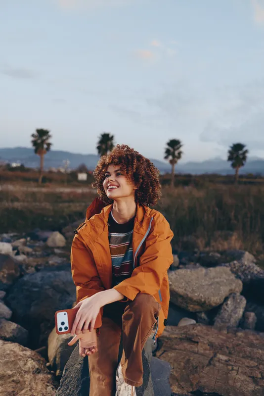 Curly hair woman lifestyle outdoors in desert landscape, orange jacket, holding smartphone, sitting on rocks, looking away, golden hour travel mood