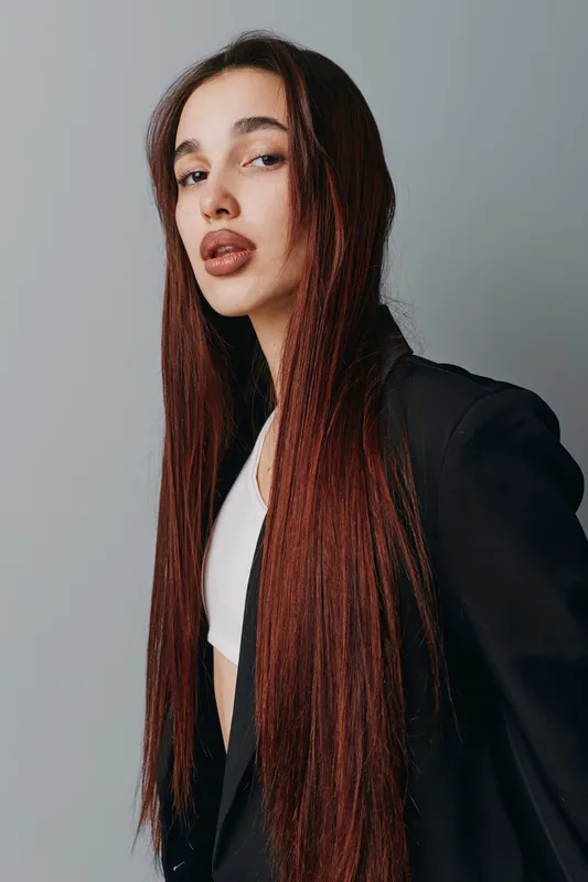 Young woman with long straight hair and makeup wearing black blazer and white top posing confidently against gray background in studio portrait fashion style.