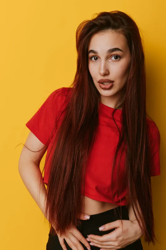 Young woman with long brown hair wearing red shirt posing against yellow background. Portrait of confident female with natural makeup and casual style in studio lighting.