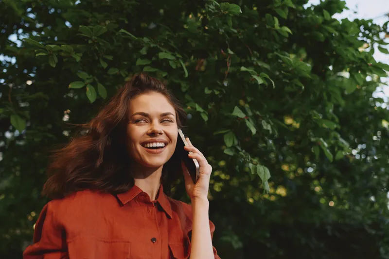 Cheerful woman outdoors, smiling while talking on a phone, sunlit garden background, casual red shirt, natural light, relaxed mood, candid lifestyle scene and warm tones overall