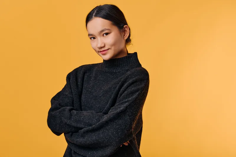 Young woman with black hair wearing dark sweater standing with crossed arms against orange background looking confidently at camera. Portrait of casual fashion and self-assurance.