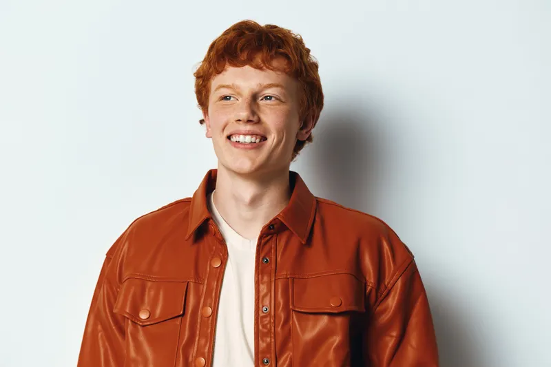 Young man with red hair and smiling face posing in brown leather jacket on light background for fashion and portrait photography. Casual style and confident expression.