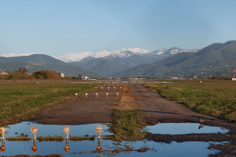 A dirt road cuts through a vast field toward distant snowcapped mountains beneath a clear blue sky, creating a tranquil, expansive rural landscape with peaceful solitude
