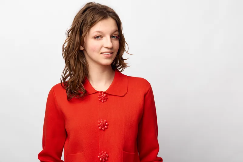 Young woman with wavy brown hair wearing a red knitted sweater with decorative buttons smiles gently against a plain white background. Casual style and natural look concept.