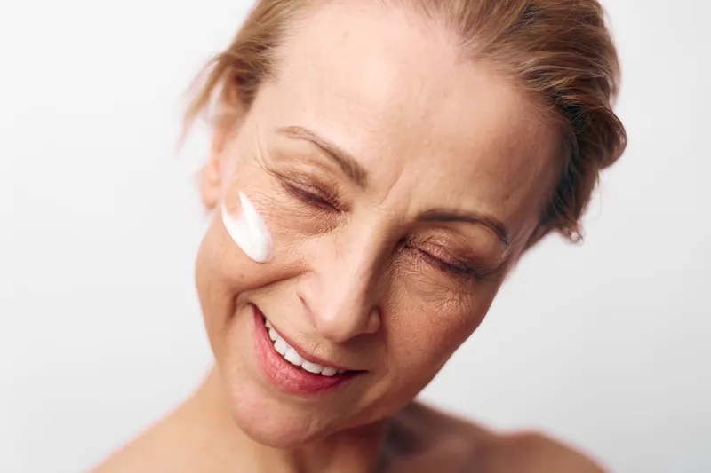 Smiling mature woman with closed eyes applies white facial cream on cheek, showing natural skin texture and self-care. Concept of skincare, wellness, anti-aging, and beauty routine.