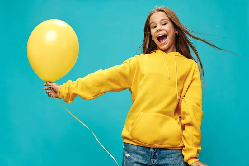 Girl smiling happily with yellow balloon wearing casual yellow hoodie and jeans on blue background for celebration and childhood fun outdoors.