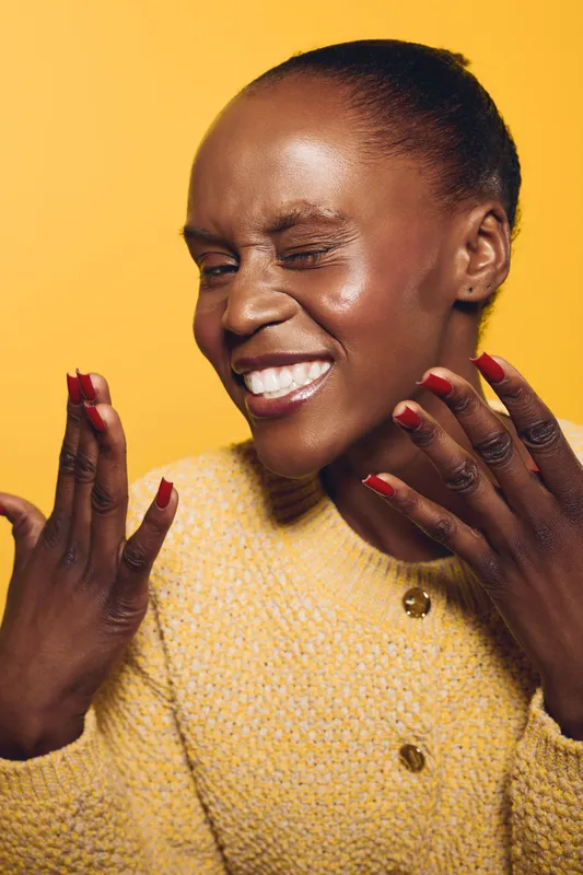 Happy African woman with dark skin wearing a yellow sweater smiles and winks with red nails on orange background. Concept of joy, beauty, and positive emotions in fashion portrait.