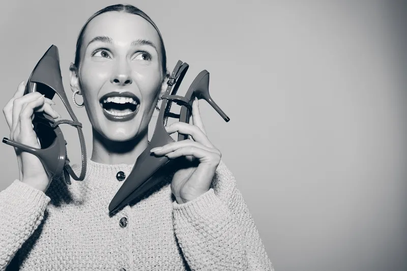 Young white woman with fair skin and dark hair holds two high heel shoes near her face, smiling with open mouth on light background. Concept for fashion, joy, lifestyle, shopping.