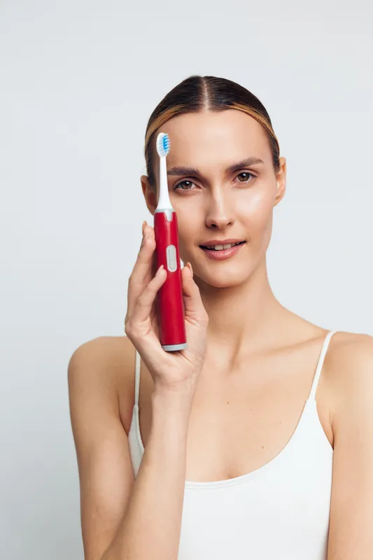Confident woman holding a red toothbrush to her face, showcasing oral care routine, clean teeth and fresh breath, isolated against a light neutral background, natural beauty