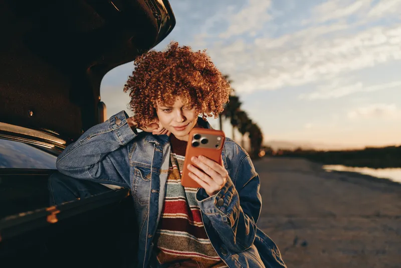 Woman lifestyle candid, curly hair woman smiles at smartphone selfie, standing by car, golden sunset on outdoor road, casual travel atmosphere, natural light street scene open sky evening