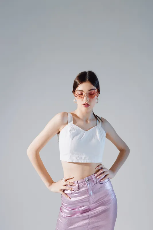 Young woman in a white crop top and shiny pink skirt poses confidently with hands on hips. Studio shot on a grey background. Concept of woman beauty fashion.