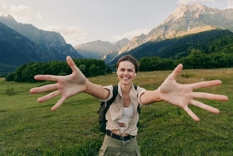 A young woman outdoors in a grassy meadow with towering mountains behind extends her arms toward the camera, smiling widely, conveying energy, freedom, and a sense of exploration.