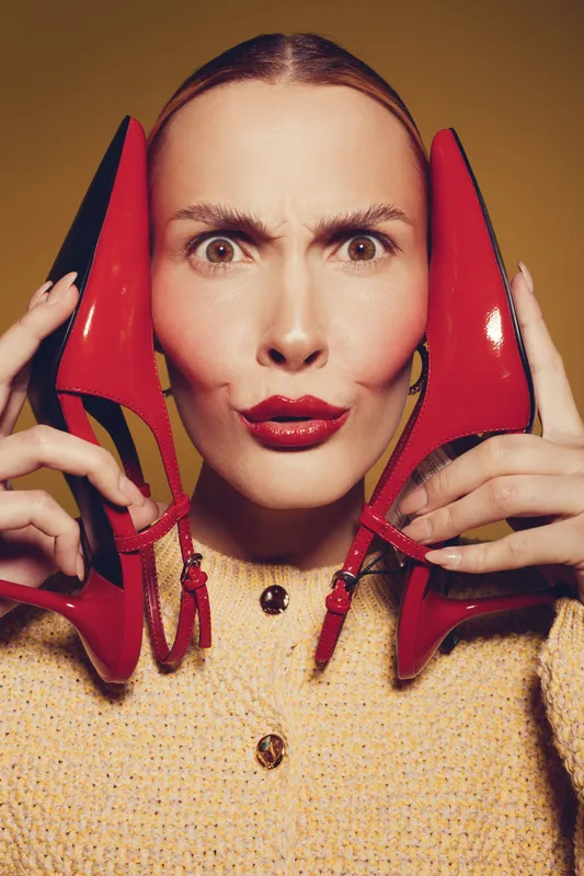 Woman with red lipstick holds red high heel shoes near face, making funny expression. Fashion concept with bright makeup and stylish accessories on beige background.
