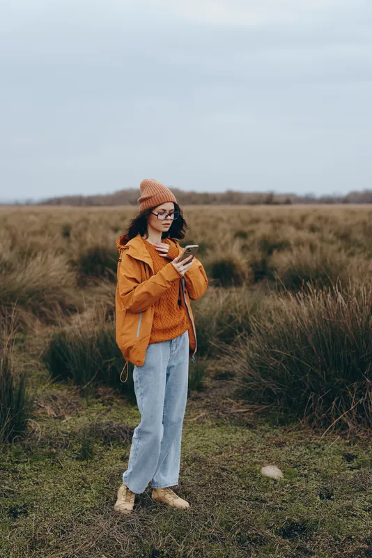 Woman outdoors in a field of tall grass, wearing brown jacket, knit beanie and denim jeans, walking in rural nature, quiet autumn lifestyle mood, natural light.
