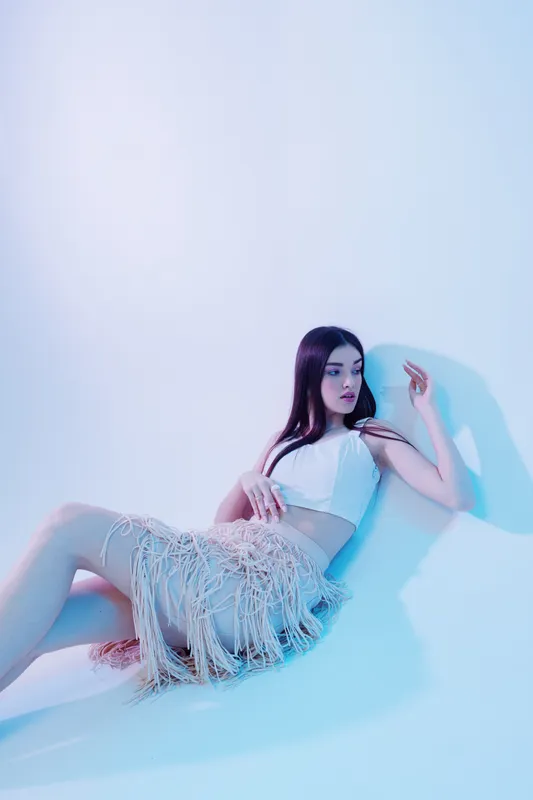 Young woman in a white top and fringed skirt poses on the floor in a studio with cool lighting. Concept of beauty, fashion, and modern style with a relaxed mood.