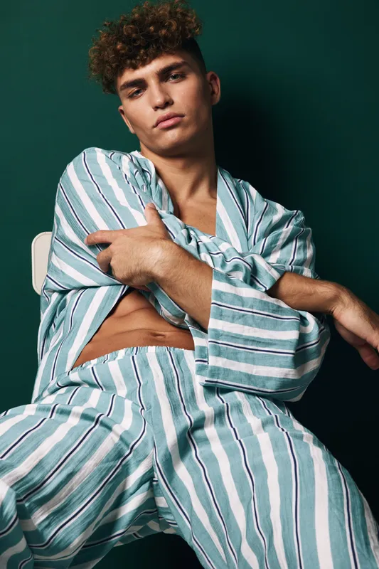 Young man with curly hair wearing striped pajamas sitting on chair with arms crossed, looking confidently at camera against dark green background in studio.