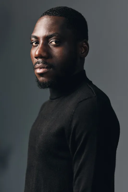 African man portrait in creative conceptual visual style wearing black turtleneck sweater with confident expression on gray background in studio lighting.