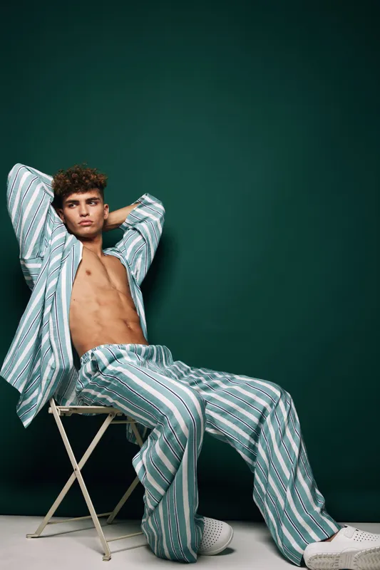 Young man with curly hair wearing striped pajamas and white shoes sitting on folding chair relaxing with hands behind head against dark green background in studio lighting.