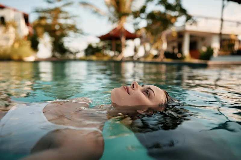 Relaxed woman floating on her back in a sunlit pool, eyes closed, enjoying calm water and a warm breeze. A tranquil lifestyle moment highlighting wellness and leisure with serenity.