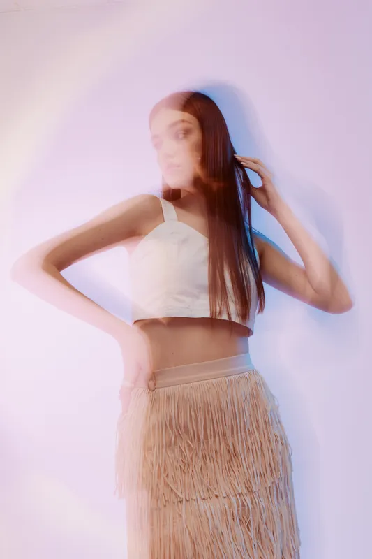 Fashionable woman with long hair wearing a white crop top and fringed skirt poses against a soft pastel background. Concept of beauty, fashion, and modern style.