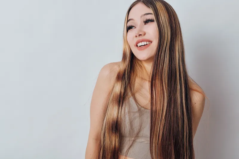 Young woman with long straight hair smiling and looking away on plain light background. Female model in casual beige top showing happiness and natural beauty style.