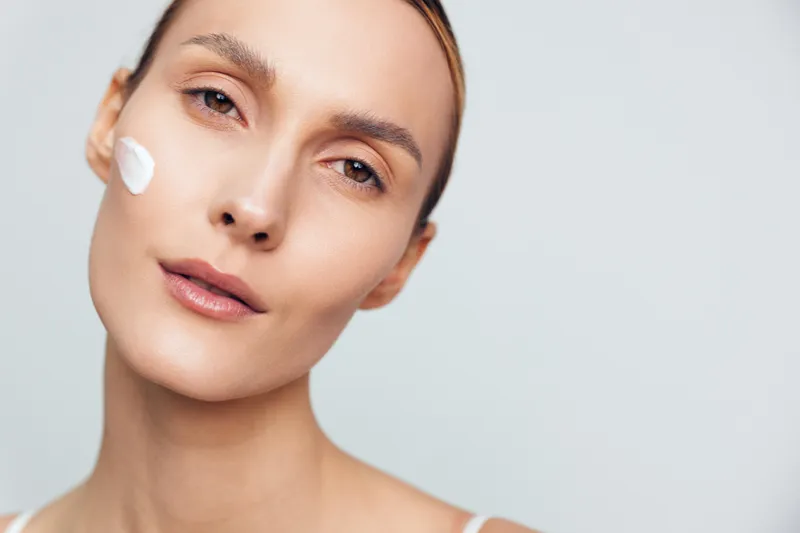 Young woman face close up, skincare beauty, cream on cheek, spa care concept, soft studio lighting on neutral background, calm expression, fresh hydration look, clean minimal portrait.