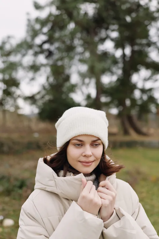Woman outdoors winter coat beanie standing in a park, looking down with a gentle smile, capturing calm, cozy outdoor style and serene mood in soft winter light.