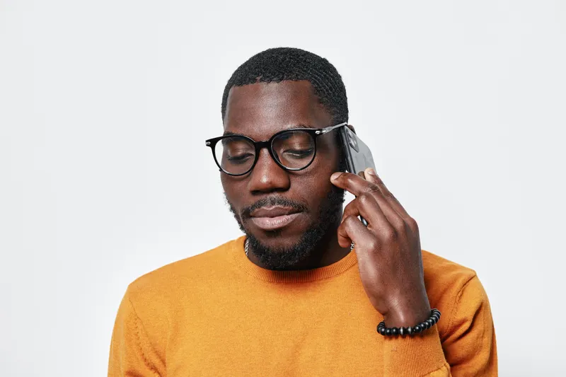 African man with dark skin and glasses talking on smartphone wearing orange sweater isolated on white background. Closeup portrait of black male using mobile phone for communication.