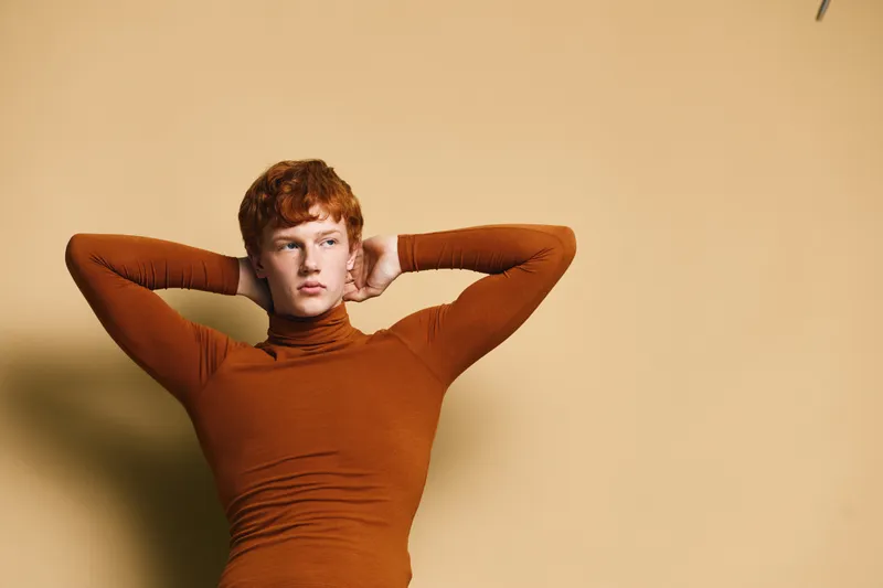 Young man with red hair wearing a brown turtleneck sweater posing with hands behind head against beige background for fashion portrait and style concept.