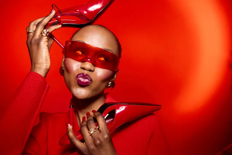 Stylish black woman with dark skin wearing red sunglasses and red outfit holds red high heels near face. Bright red background creates bold fashion and beauty concept with confident mood.