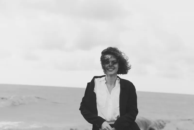 Woman on a windy beach wearing a white shirt and dark jacket, smiling against a cloudy sky, enjoying a breezy outdoor seaside moment with relaxed style